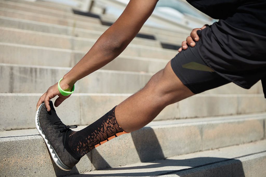 Cropped shot of male runner in black sportswear stretching legs before doing morning workout. Young African jogger warming up before outdoor run, standing on steps of concrete stair, touching toes